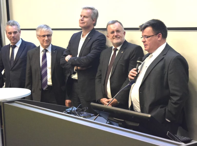 Fünf Männer in Anzügen bei einer Veranstaltung, einer spricht am Mikrofon neben einem Rednerpult