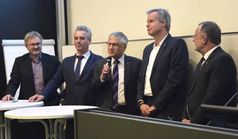 Fünf Männer in Anzügen stehen bei einer Veranstaltung, einer spricht ins Mikrofon