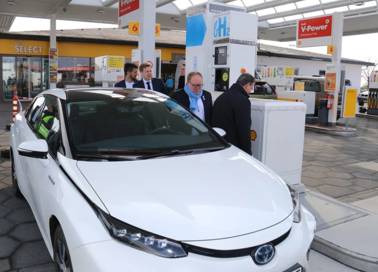 Männer in Jacken begutachten ein weißes Wasserstoffauto an der H2-Zapfsäule einer Tankstelle