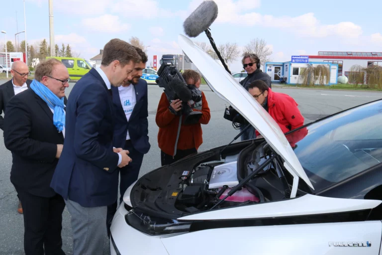Mehrere Personen begutachten den geöffneten Motorraum eines Wasserstoff-Fahrzeugs, Kamerateam filmt die Szene