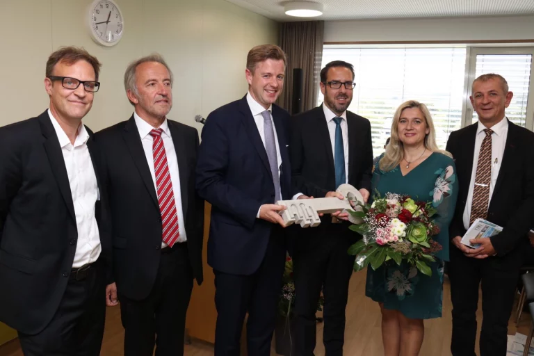 Gruppe von Personen übergibt Blumenstrauß und Modell des Klinikneubaus in einem Büroraum