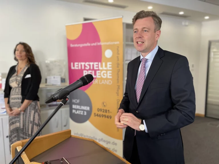 Mann im Anzug spricht am Rednerpult mit Banner der Leitstelle Pflege Hofer Land, Frau daneben