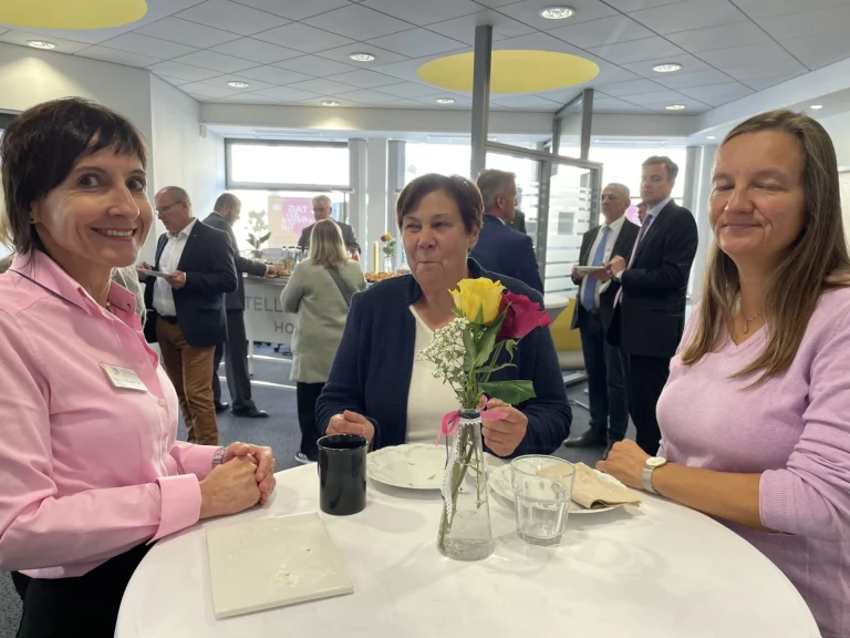 Frau mit Blumenstrauß am Tisch im Gespräch, im Hintergrund Gäste bei einem Stehempfang