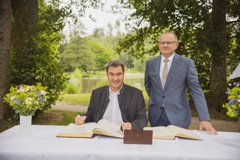Markus Söder trägt sich bei Unterzeichnung im Garten in ein Gästebuch ein, daneben ein Mann im Anzug