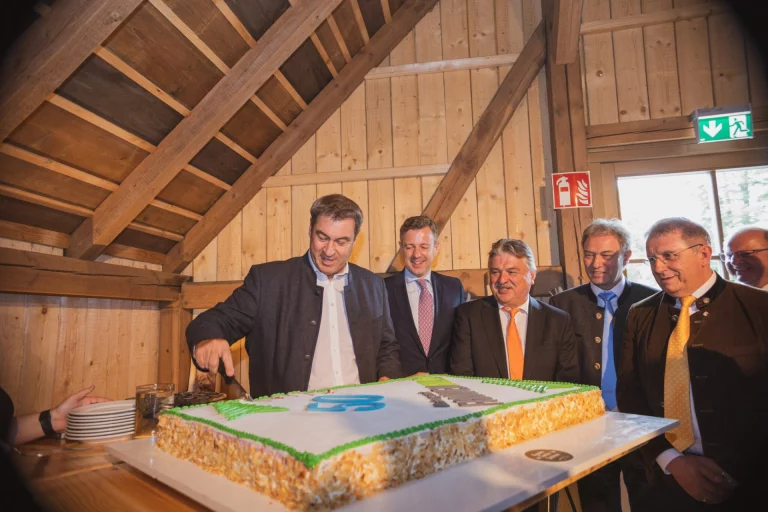 Markus Söder schneidet Jubiläumstorte an, umgeben von Vertretern bei Festakt im Holzhaus