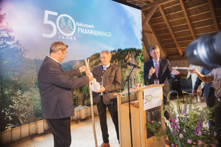 Männer bei Übergabe eines Wanderstocks vor Bühnenwand „50 Jahre Naturpark Frankenwald“