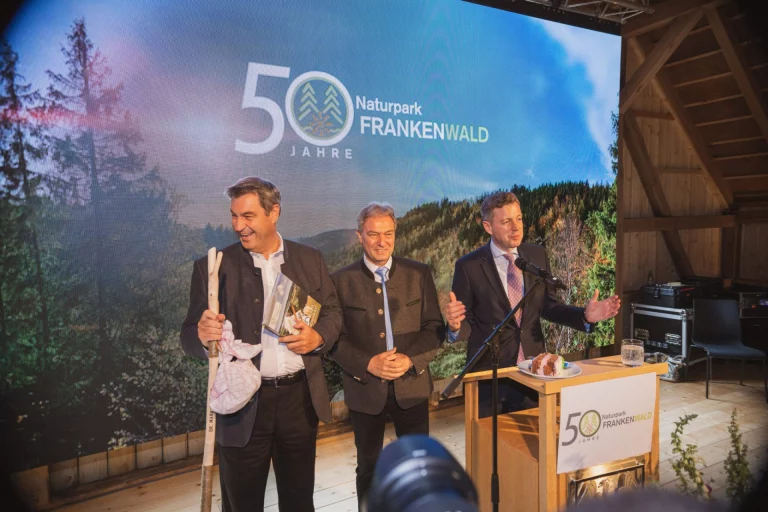 Drei Männer auf einer Bühne vor Leinwand „50 Jahre Naturpark Frankenwald“ am Rednerpult