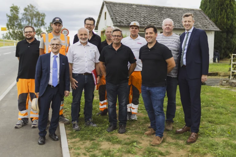 Gruppe von Personen in Arbeitskleidung und Anzug an der neuen Kreisstraße nahe einem Bauernhaus
