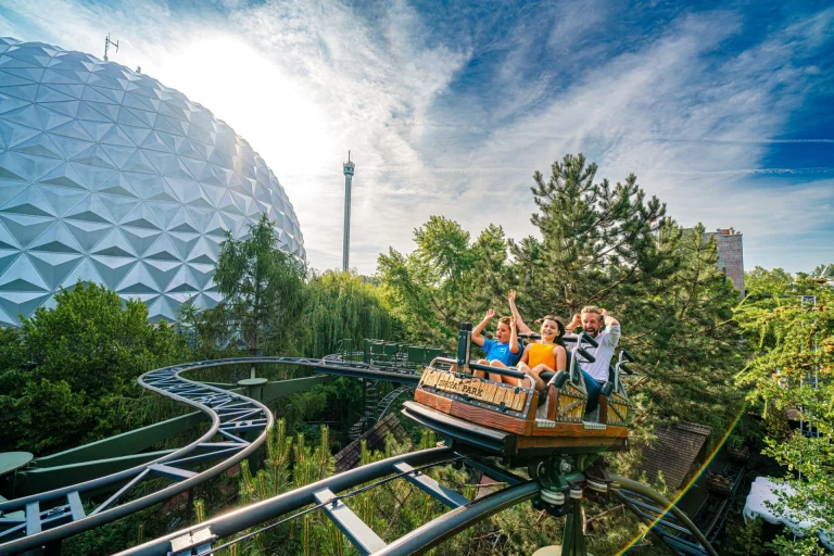 Kinder und ein Mann jubeln bei einer Achterbahnfahrt im Freizeitpark vor einer Kuppelbau-Attraktion