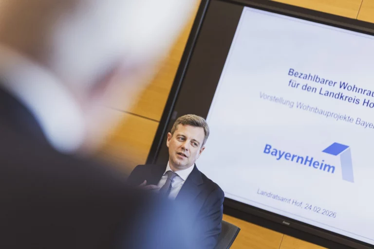 Mann im Anzug im Gespräch, im Hintergrund Bildschirm mit BayernHeim-Präsentation zu bezahlbarem Wohnraum