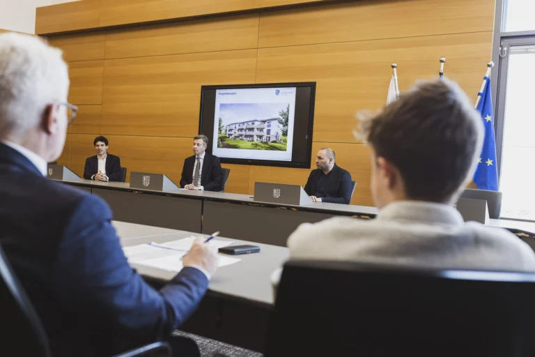 Männer an Konferenztisch, im Hintergrund Bildschirm mit Visualisierung eines Wohngebäudes