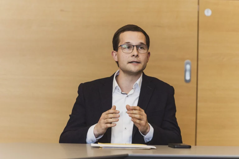 Mann mit Brille im Anzug spricht sitzend am Tisch, Notizblock und Smartphone liegen bereit
