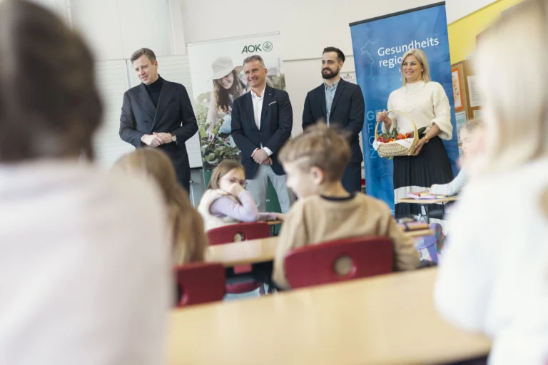 Erwachsene und Grundschulkinder in Klassenzimmer, Frau mit Gemüsekorb vor Rollup „Gesundheitsregion“