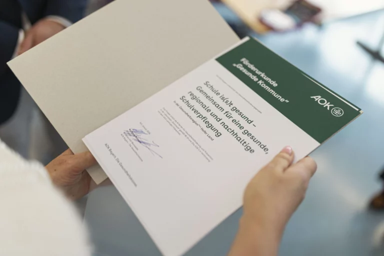 Hände halten Förderurkunde „Gesunde Kommune“ der AOK zum Projekt Schule is(s)t gesund