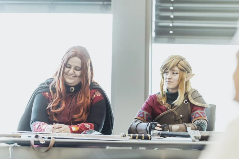 Zwei Personen in Fantasy-Cosplay mit Umhang und Schwertern an einem Tisch