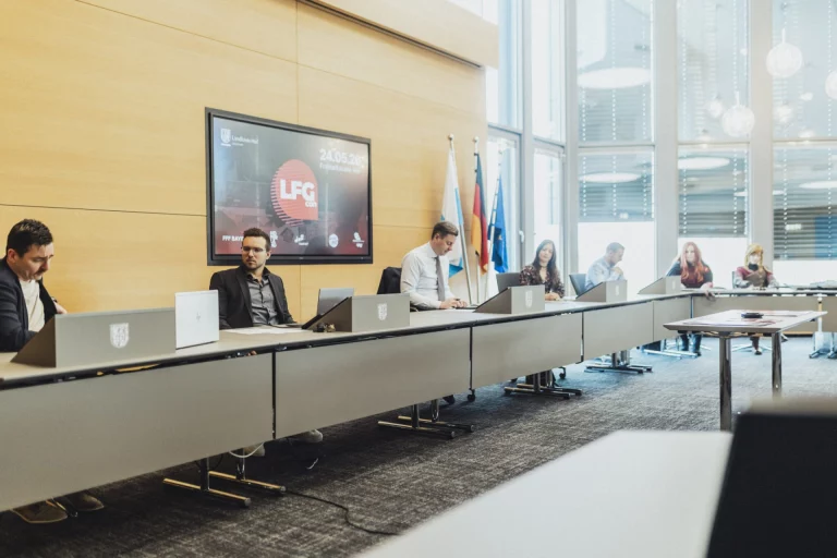 Personen mit Laptops an Tischreihe im Sitzungssaal, Bildschirm zeigt LFG-Logo