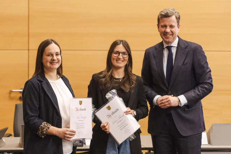 Zwei Frauen mit Urkunden stehen neben einem Mann im Anzug bei einer Ehrung