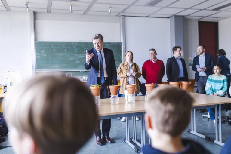 Mann im Anzug spricht zu Schulklasse an Tisch mit Pflanztöpfen und Wasserflaschen im Klassenzimmer