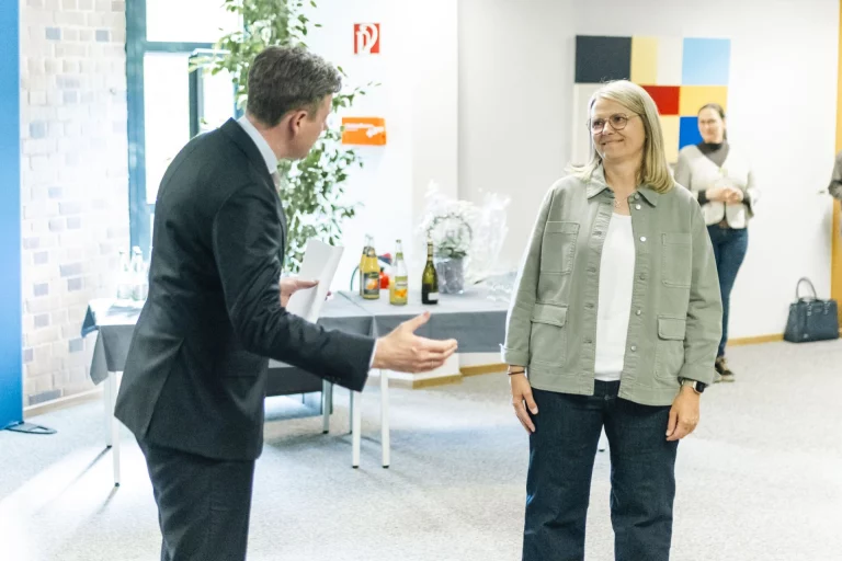 Mann im Anzug begrüßt lächelnde Frau in grüner Jacke bei der Ausstellungseröffnung