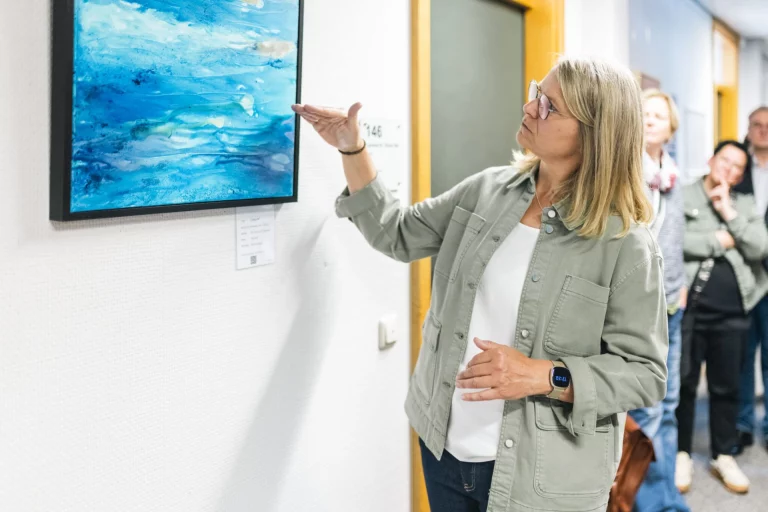 Frau erläutert ein abstraktes blaues Acrylbild, im Hintergrund weitere Besucher der Ausstellung