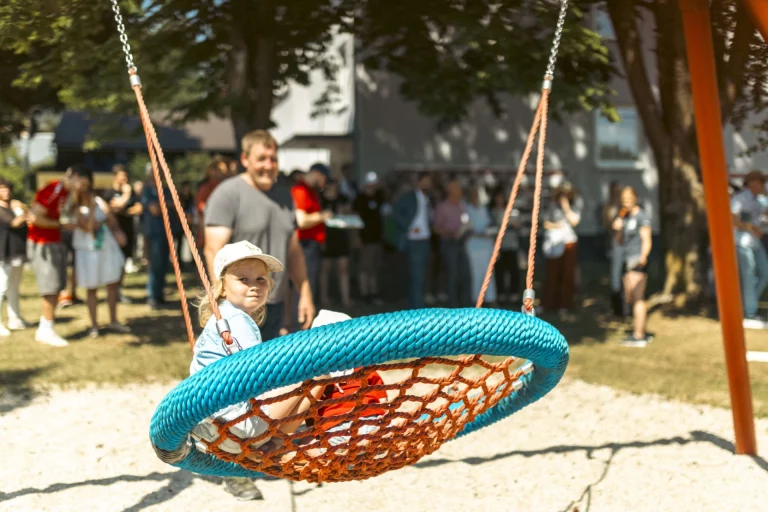 Kind in Nestschaukel auf Spielplatz, im Hintergrund Besuchergruppe bei Dorffest in Hallerstein